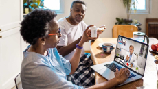 Person on laptop at kitchen table with another who holds up prescription bottle for telehealth doctor