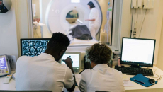Radiologists look on screen as a patient exiting an MRI machine appears in the background.
