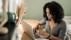 A person wearing a digital health-tracker watch sitting at a desk by a couch types on a laptop.