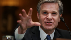 FBI Director Christopher Wray reaches his hand forward as he speaks into a microphone.