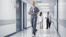 Nurses and doctors walk down a hospital hallway, one checks patient monitoring data on a tablet.