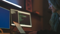 Person with long hair sits before a laptop and multiple screens