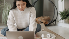 A woman with long hair sits on her floor at home as she engages in a behavioral health appointment.
