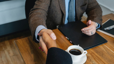 Seated individuals are shaking hands over a table and a cup of coffee.