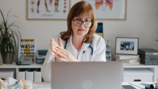 A doctor with glasses sits at a desk and speaks during a telehealth visit.