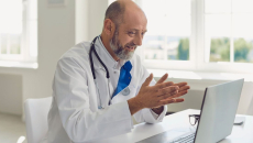 Healthcare provider with a lab coat and stethoscope sitting at a desk speaking with someone on a computer
