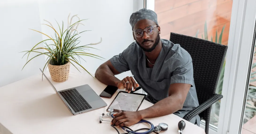 Doctor in scrubs sits at a desk near a patio with stethescope, tablet, laptop and phone.