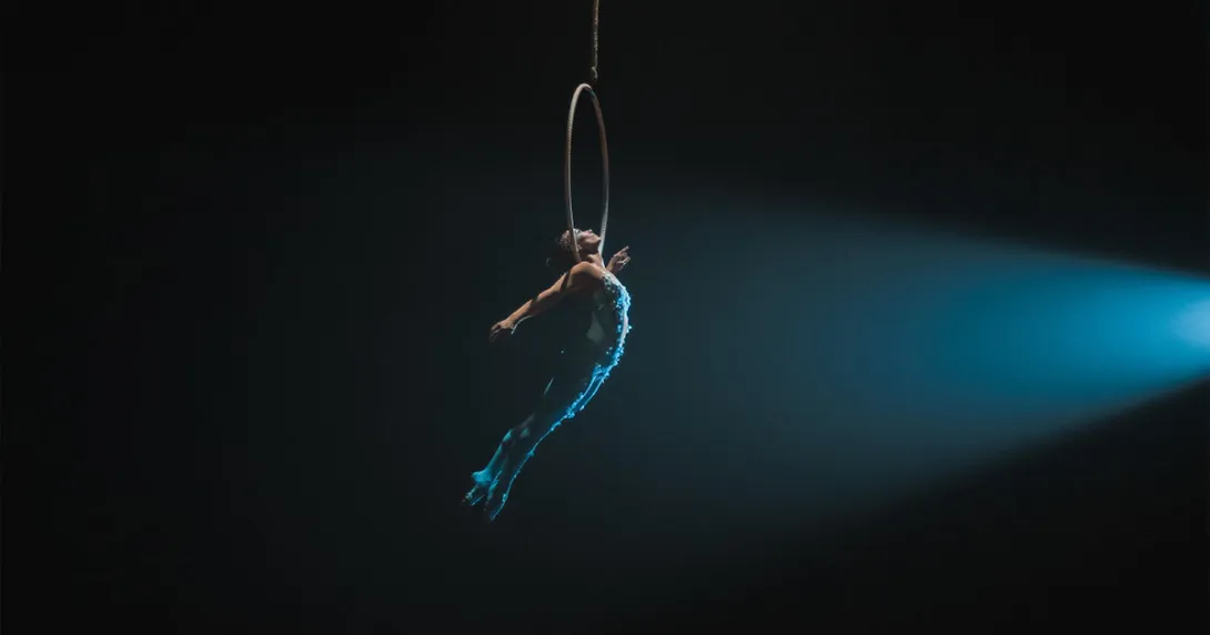 Trapeze artist in a blue costume holding onto a hoop in mid-air against a black backdrop