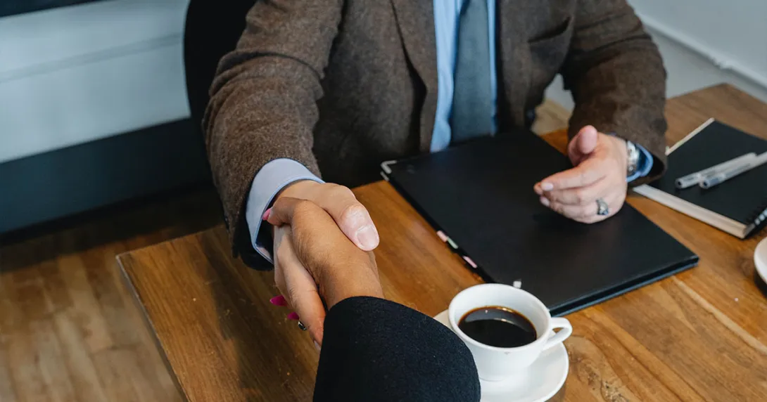 Seated individuals are shaking hands over a table and a cup of coffee.