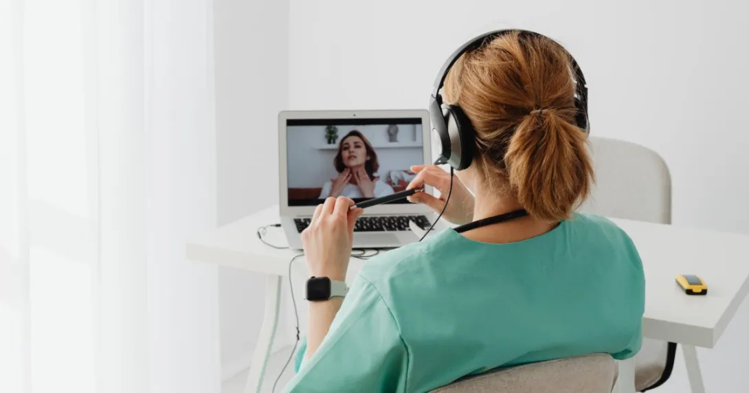A clinician in green scrubs with red hair is conducting a virtual visit with a patient.