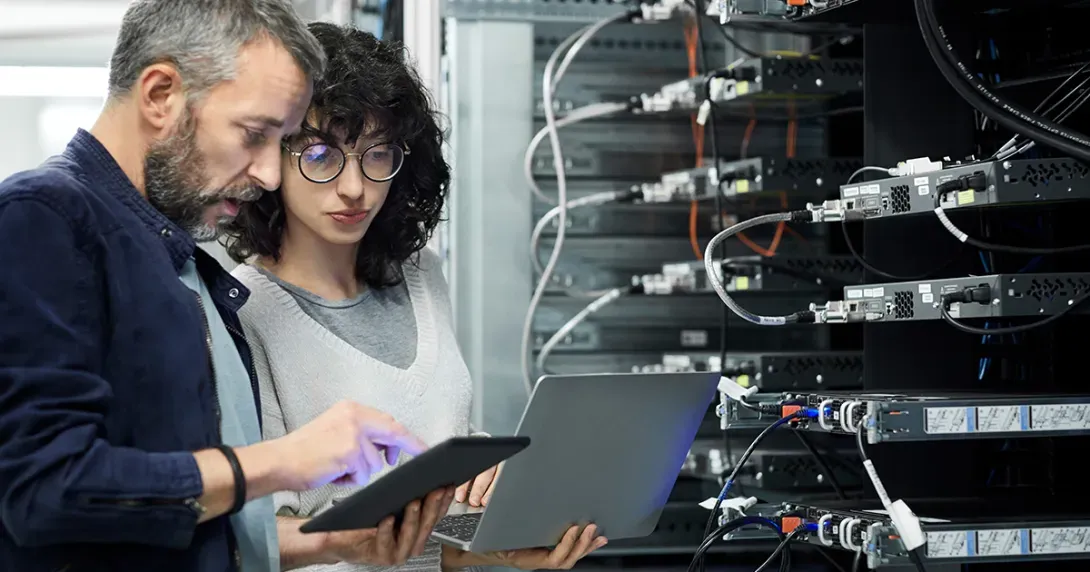 IT staff in a server room look at data on a laptop and tablet 