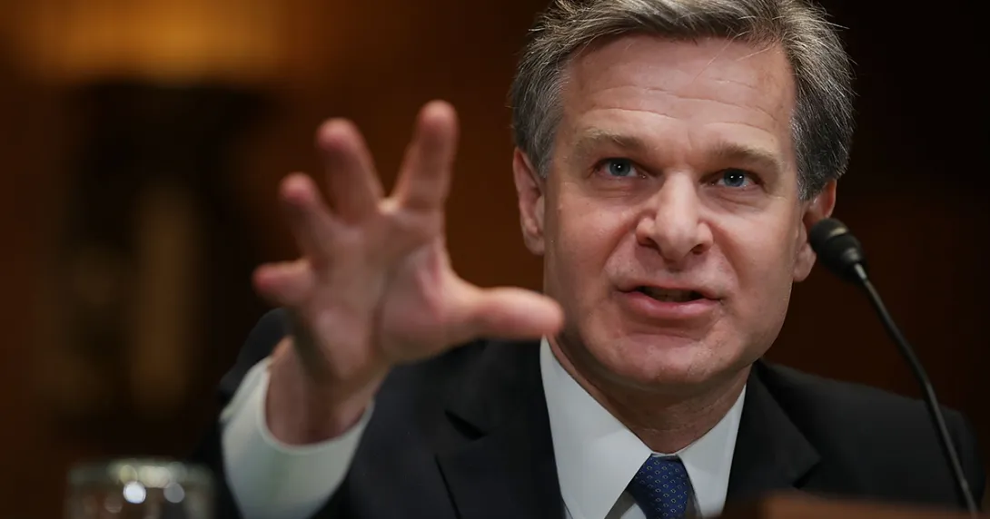 FBI Director Christopher Wray reaches his hand forward as he speaks into a microphone.