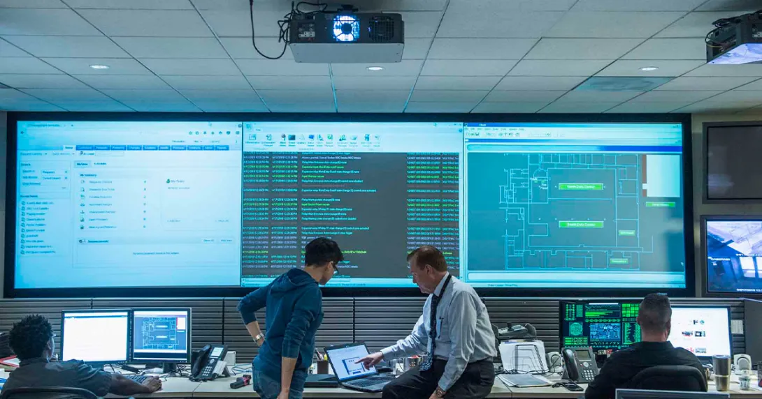 Workers looking at a laptop with computer code displayed on a big screen behind them