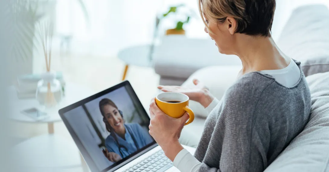 Person sitting on a couch speaking to a healthcare professional on a computer while holding a cup in their hand