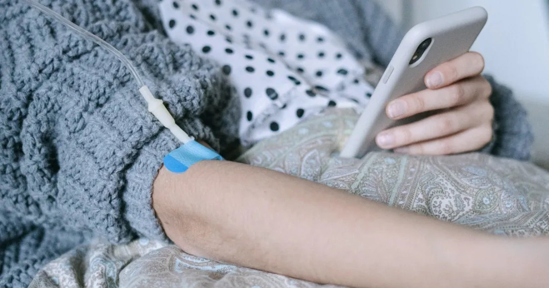 Patient in a blue sweater comforter recuperates at home and receives an IV infusion while checking patient portal information on a cell phone.