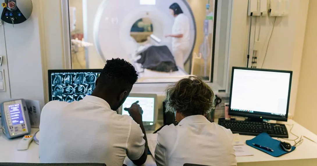 Radiologists look on screen as a patient exiting an MRI machine appears in the background.