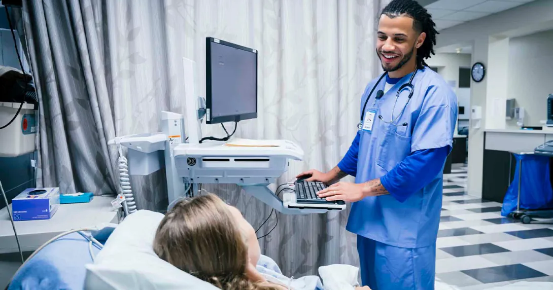 Healthcare worker at computer talking to patient in hospital bed