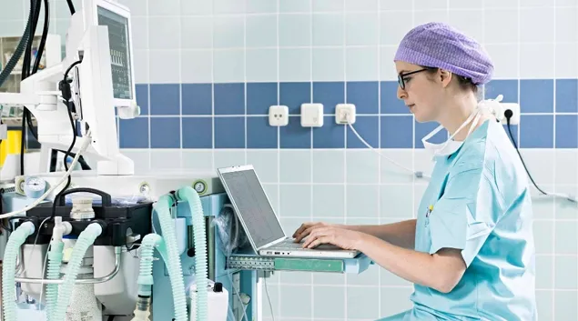 Healthcare worker in scrubs using laptop next to healthcare machinery