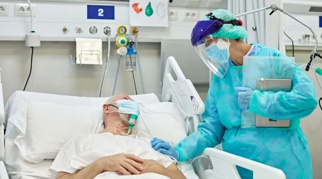 Doctor in scrubs and PPE with hand on masked patient in hospital bed