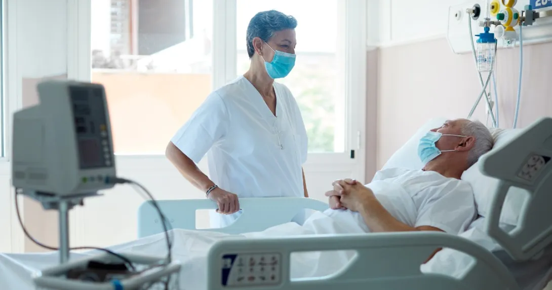A hospital staff member stands at a patient's bedside.