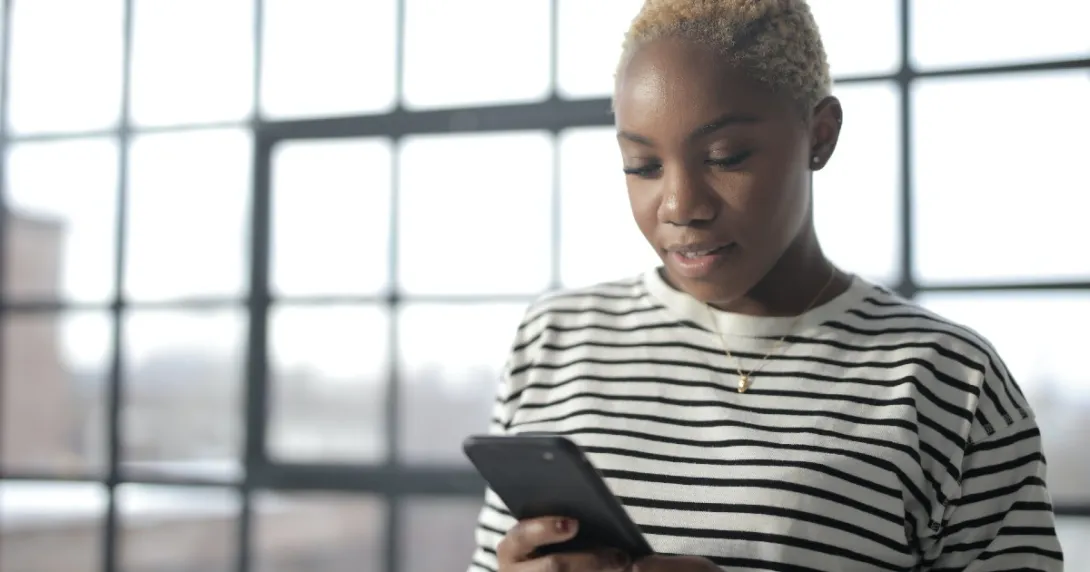 A woman of color with a striped shirt looks happily at her mobile phone.
