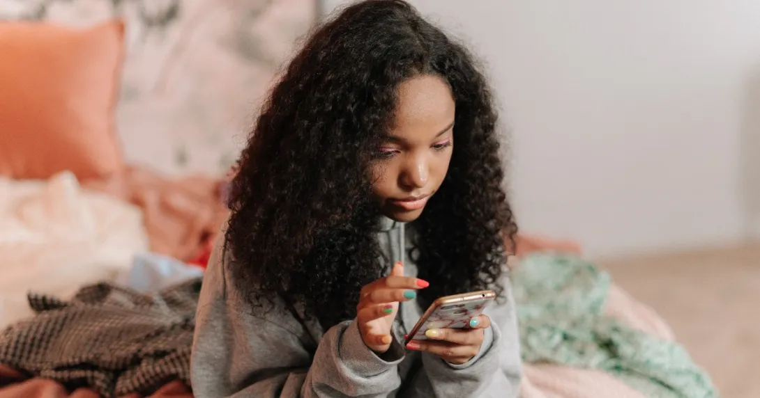 Young woman wearing a sweatshirt is sitting on her bed and checking a mobile application for information.