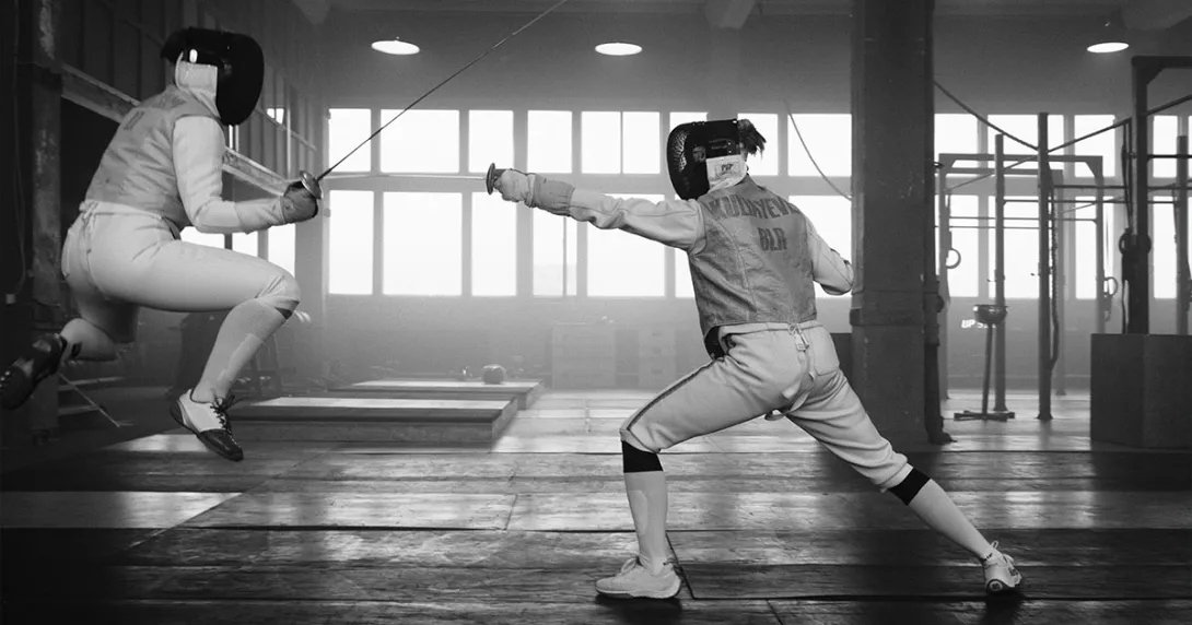 Black and white image of two individuals fencing. One is jumping but the opponent finds the weak spot.