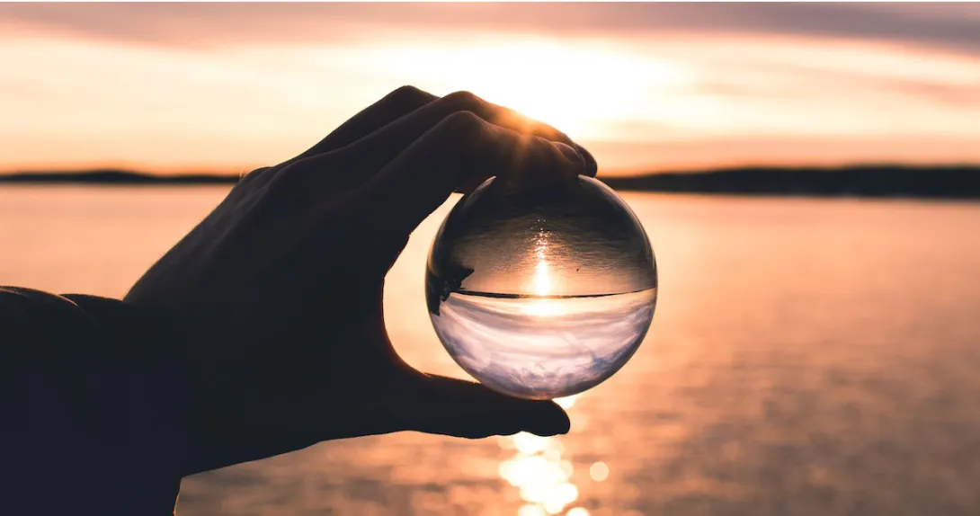 Hand holds a crystal ball before a sunset scene that is reflected inside the ball.