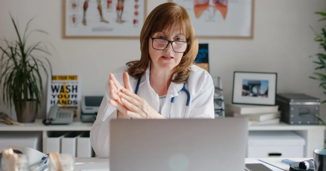 A doctor with glasses sits at a desk and speaks during a telehealth visit.
