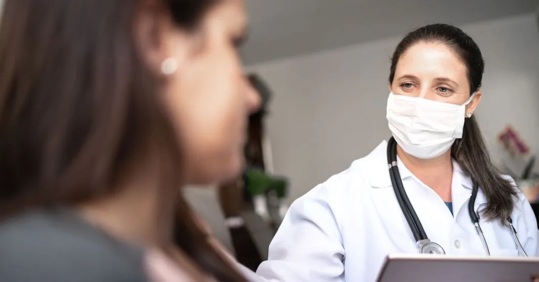 A doctor wearing a face mask and holding a digital tablet is in a face-to-face consultation with a patient