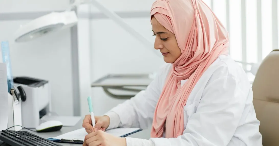 A hijab-wearing nurse filling out a patient form on a desk