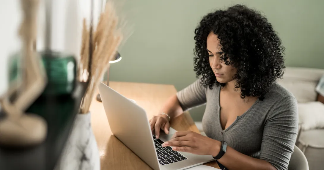 A person wearing a digital health-tracker watch sitting at a desk by a couch types on a laptop.